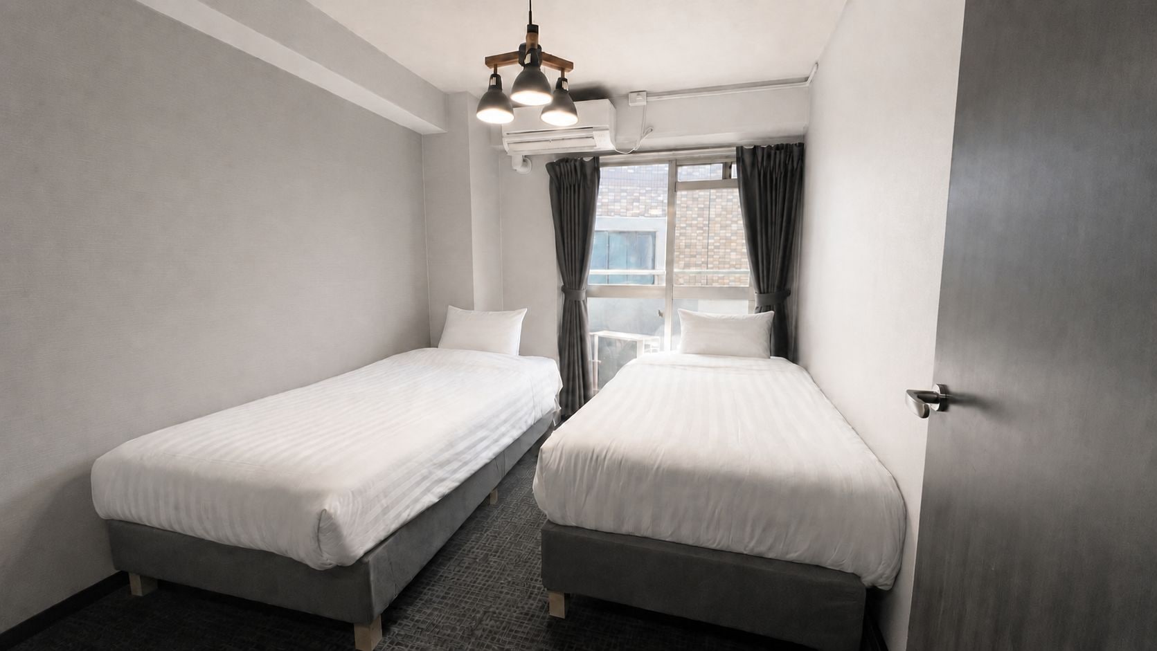 A hotel room with two single beds, a window with curtains, a ceiling light fixture, and a door.