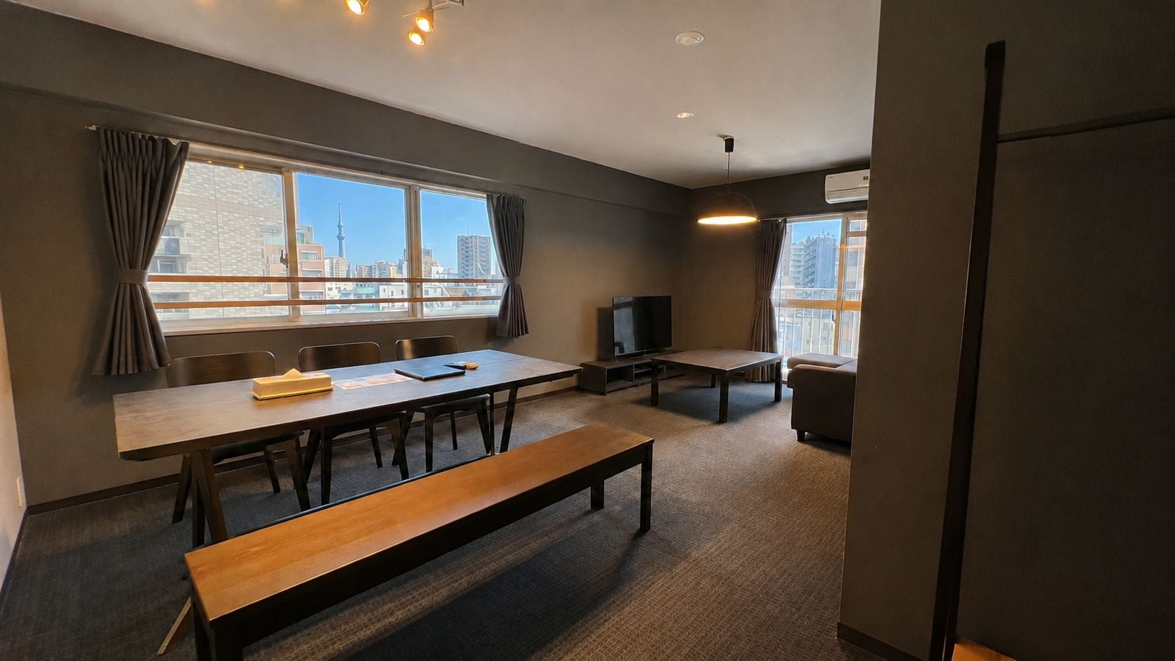 A conference room with a long table, chairs, a bench, a television, and a city view through large windows.