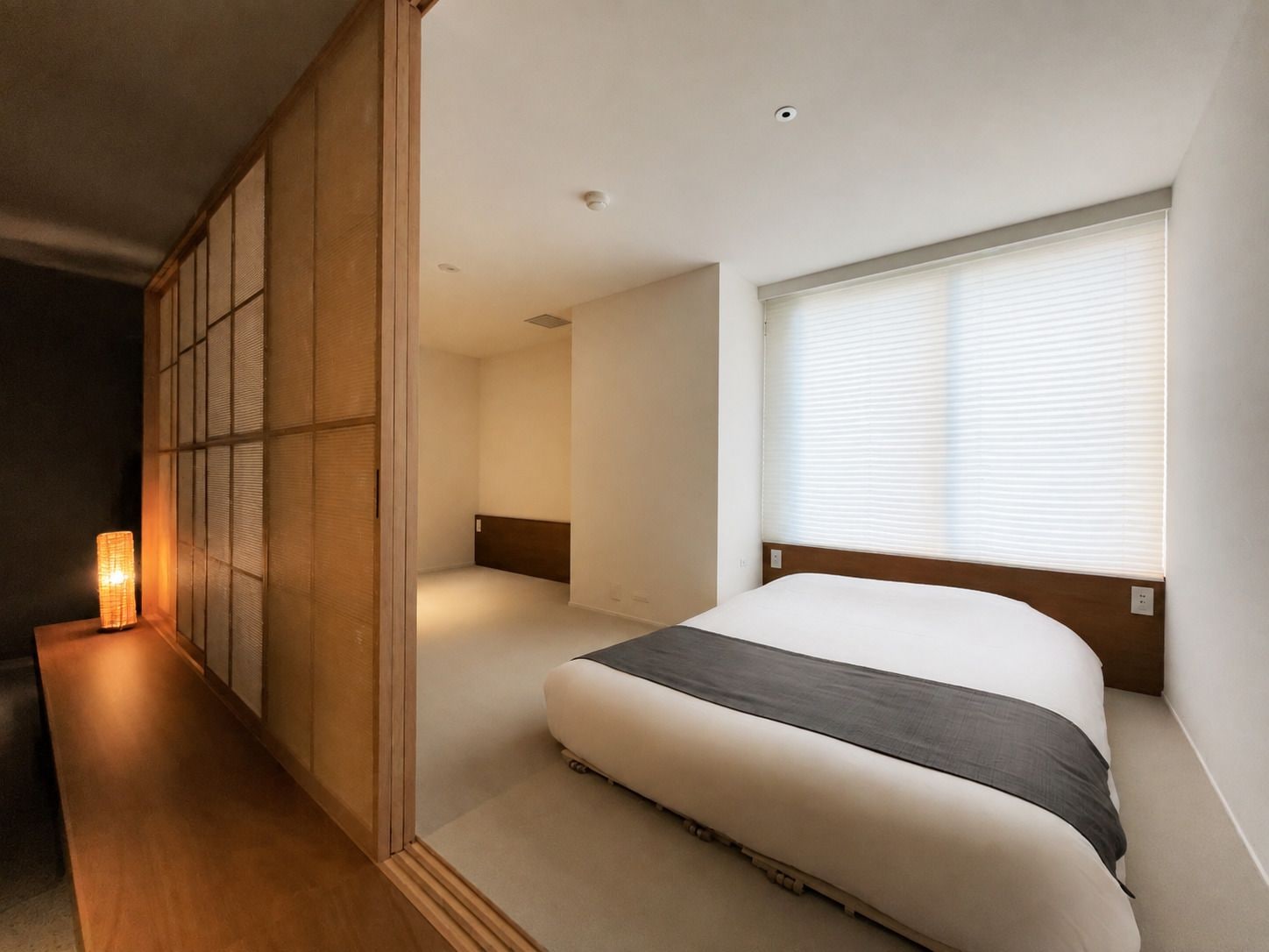 A bedroom with a bed, a window with blinds, a bedside table with a lamp, and a sliding door.