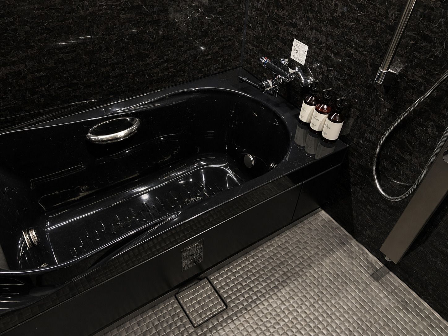 Una bañera negra con un cabezal de ducha manual, un panel de control y tres botellas de champú y acondicionador.