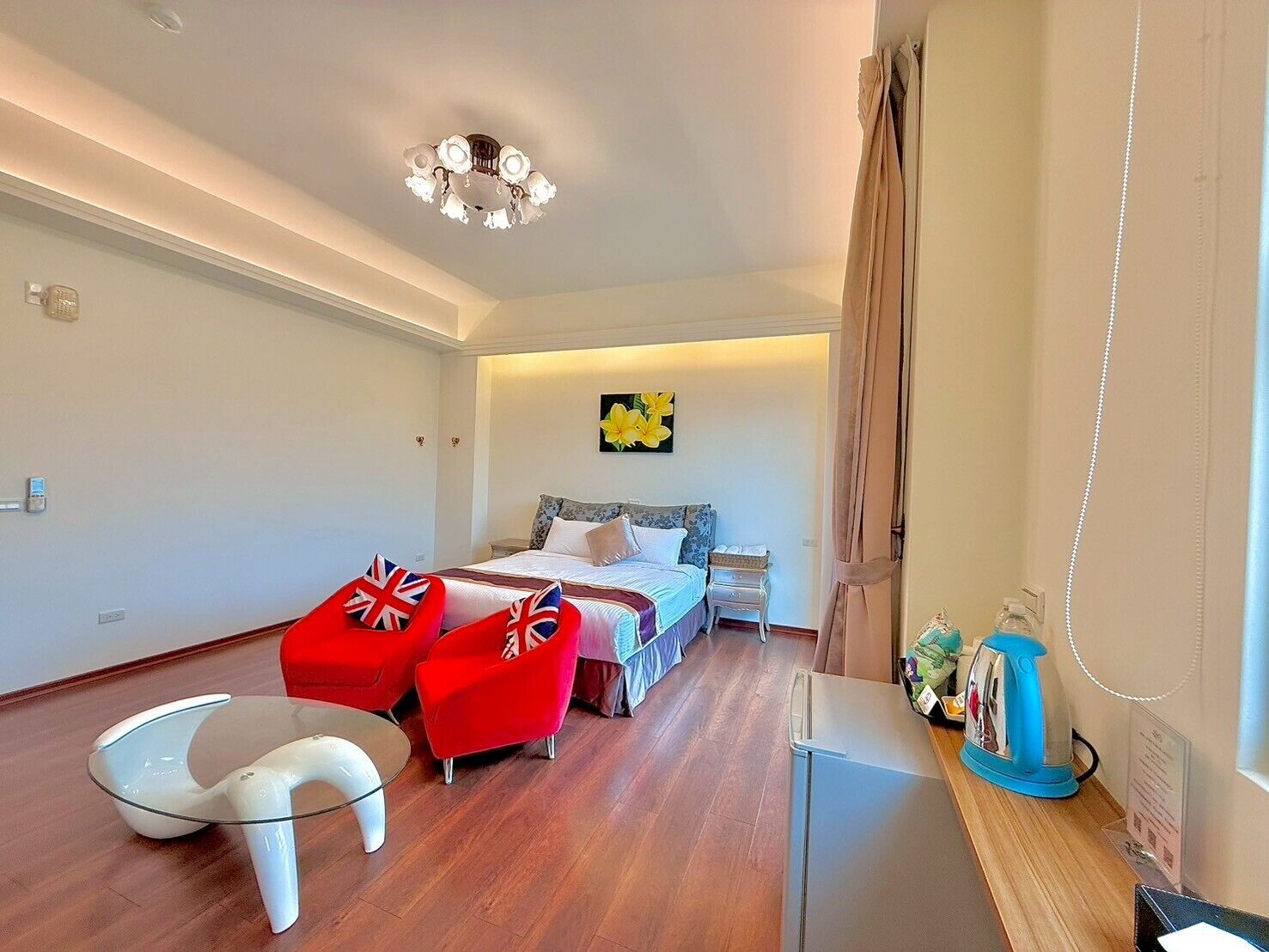 Una habitación de hotel moderna con una cama, dos sillones rojos, una mesa de centro de cristal, un escritorio con hervidor y una lámpara en el techo.