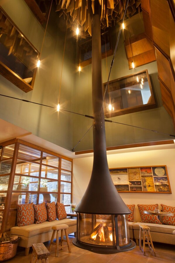 A cozy living room with a central fireplace, wooden beams, and a high ceiling.