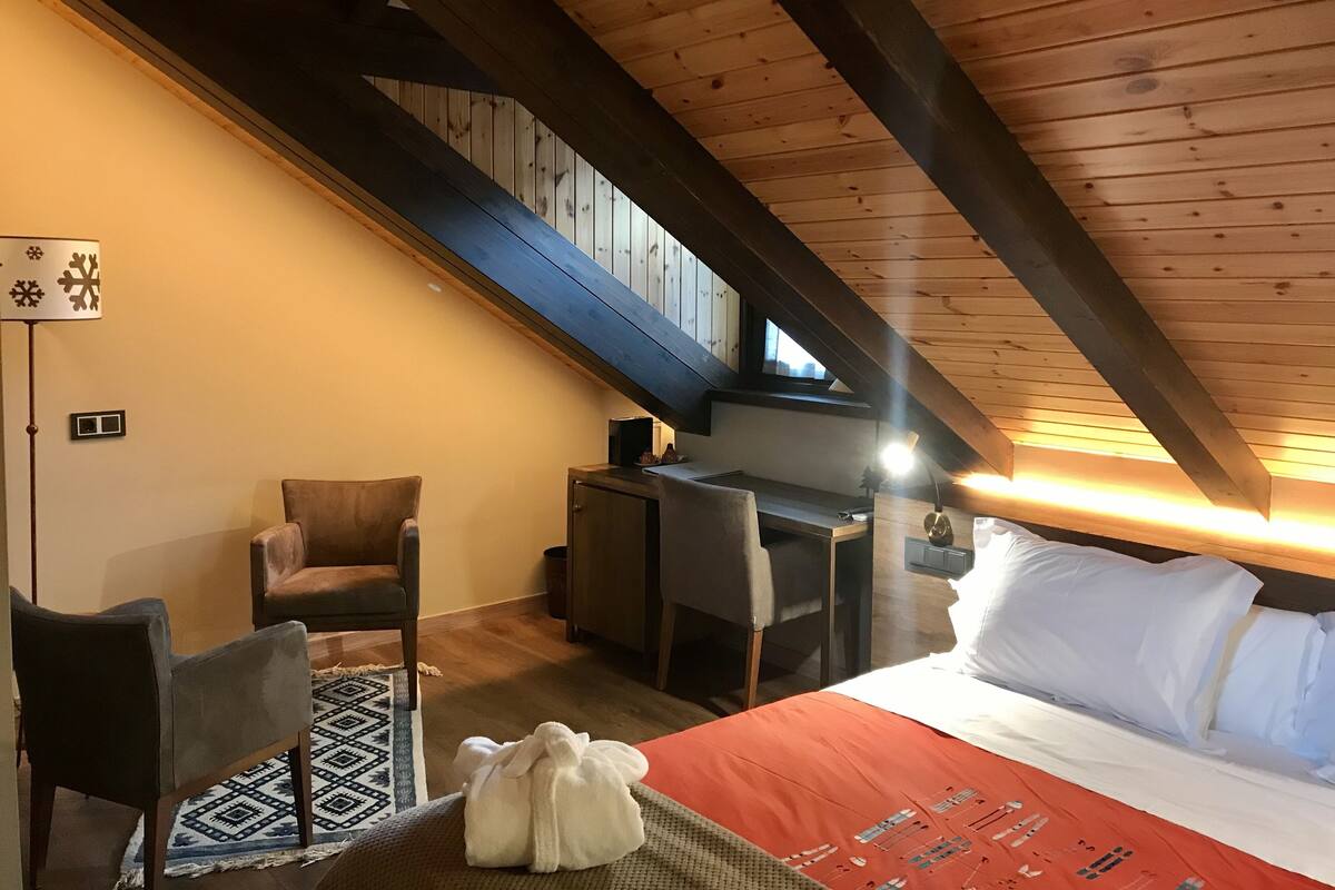 Un dormitorio acogedor en el ático con techo inclinado, una cama con ropa blanca, un escritorio con silla y una alfombra estampada.