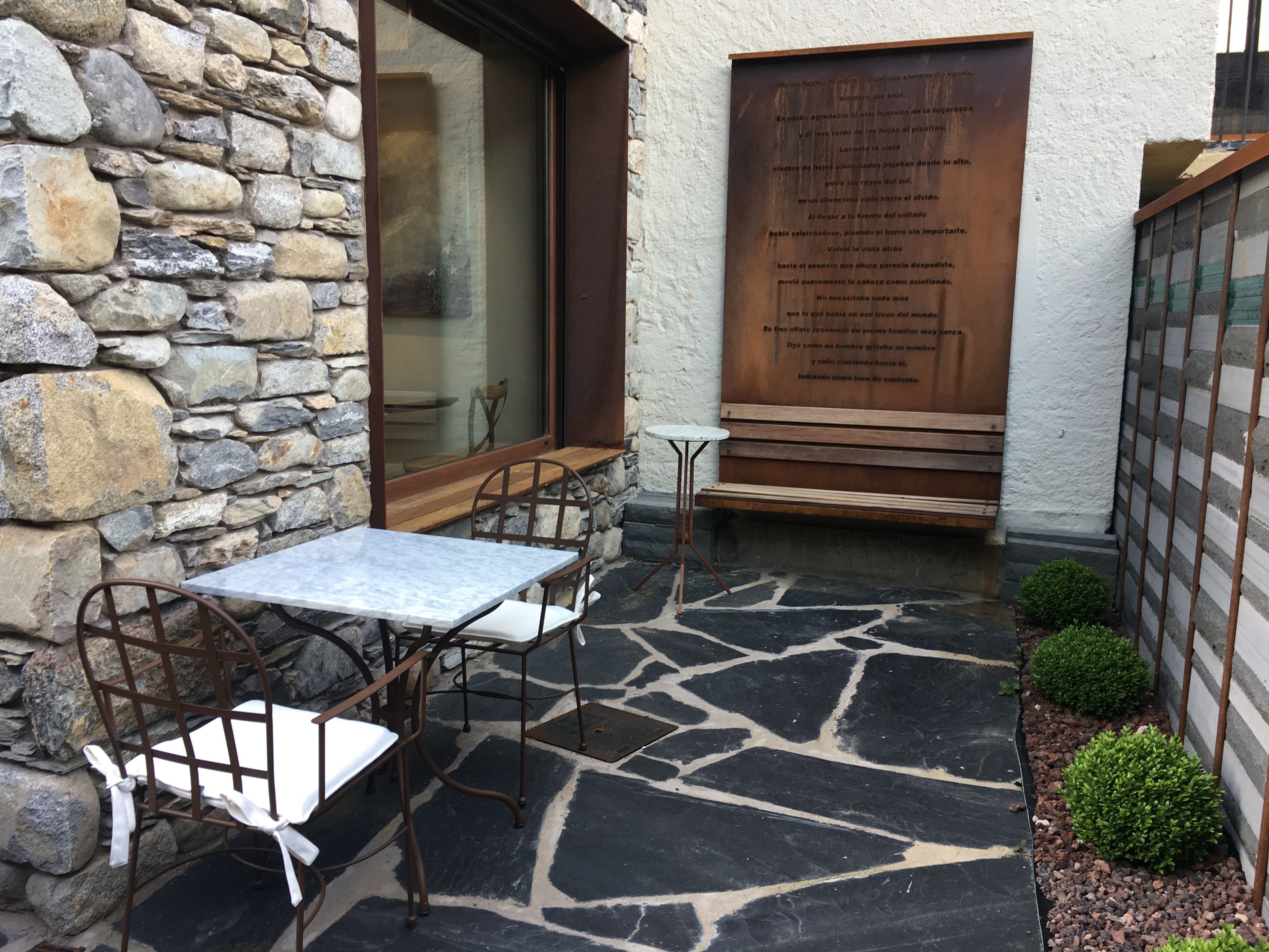 A stone wall, a wooden door with a plaque, a table with chairs, and a small garden area.