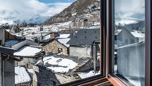 Une vue depuis une fenêtre donnant sur un village enneigé, avec des bâtiments traditionnels et des montagnes en arrière-plan.