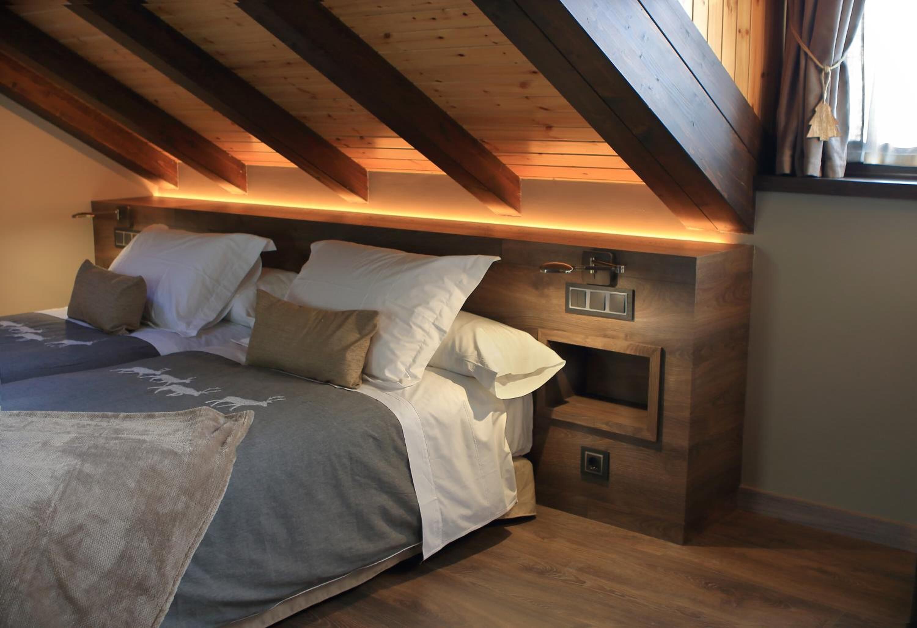 Un dormitorio con techo de madera, una cama con muchos almohadones, una mesita de noche y otra mesita auxiliar.