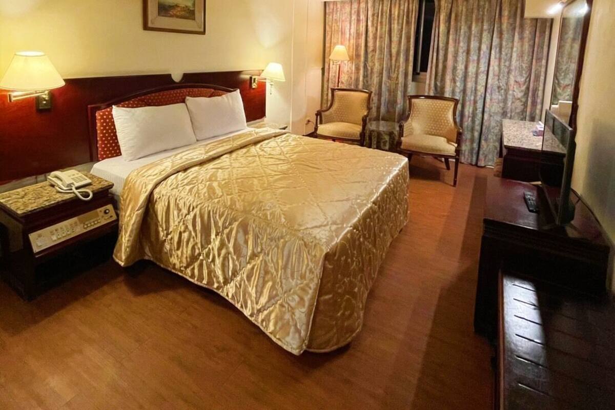 A hotel room with a large bed, two bedside lamps, a telephone, a desk with a lamp, a chair, and a television.