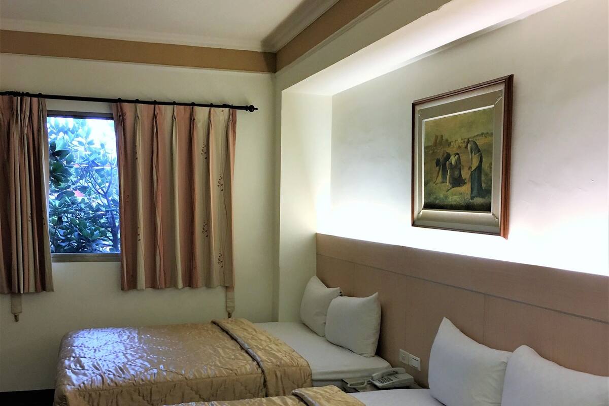 A hotel room with two beds, a window with curtains, a painting on the wall, and a telephone on the nightstand.