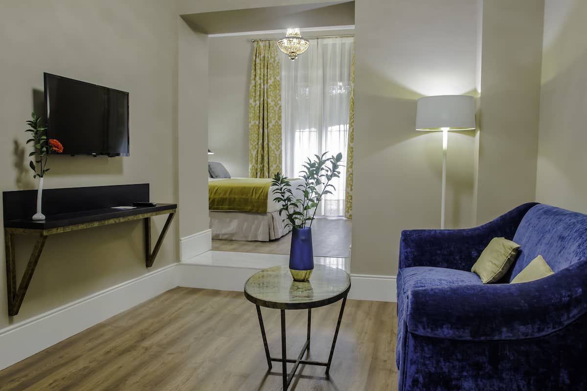 A living room with a blue sofa, a small round table with a vase, a TV mounted on the wall, and a bed with a yellow blanket in the background.