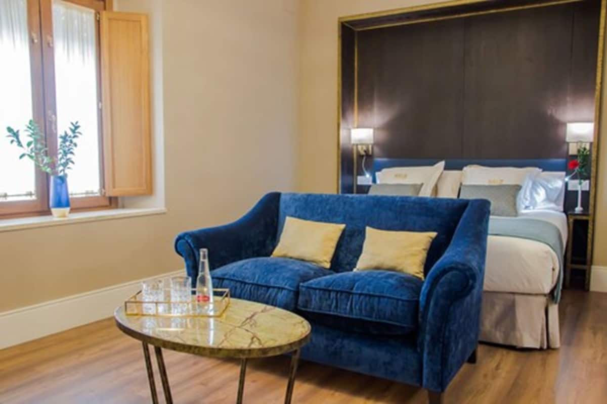 A room with a blue sofa, a small round table with glasses, a bed with white bedding, and a window with curtains.
