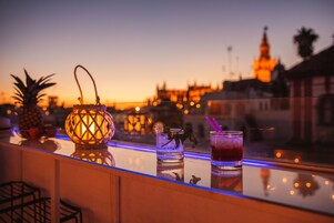 Un bar con bebidas y un farol en un balcón con vistas a un paisaje urbano al atardecer.