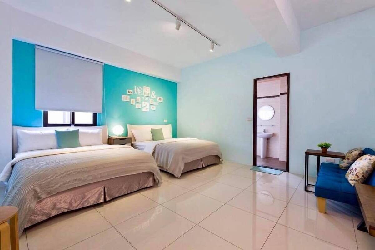 A modern hotel room with two beds, a blue accent wall, a small seating area, and a visible bathroom.