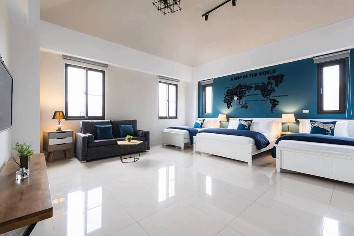 A modern, well-lit room with a large world map mural, a grey sofa, two white beds, and a wooden table with a lamp.