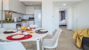 A modern kitchen with white cabinets, a dining area with a white table set for two, and a visible bed in the adjacent room. - SAS Appart Hotel Mandelieu (Mandelieu la Napoule)