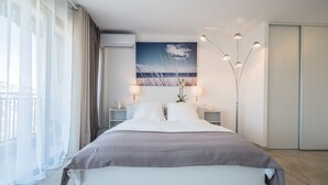 A modern hotel room with a large bed, a view of the beach, a ceiling fan, and a floor lamp. - SAS Appart Hotel Mandelieu (Mandelieu la Napoule)