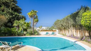 A swimming pool with lounge chairs, surrounded by palm trees and other greenery, with a clear view of the ocean in the background. - Casa da Montanha (Lagos)