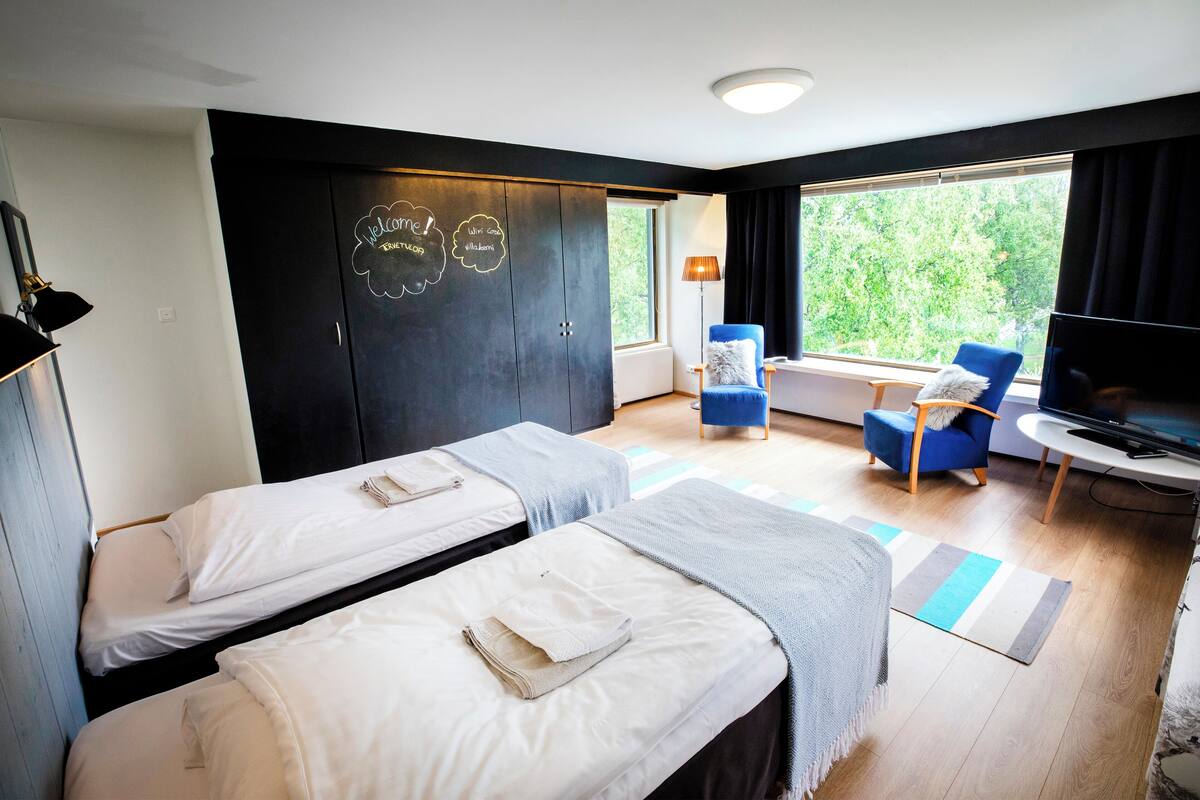 Una habitación con dos camas, televisión, una silla y ventana con vistas a la vegetación.