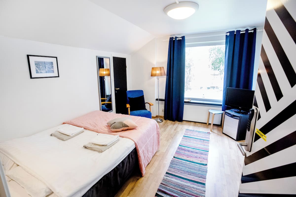 Una habitación de hotel con cama, escritorio, silla, lámpara y ventana con cortinas.