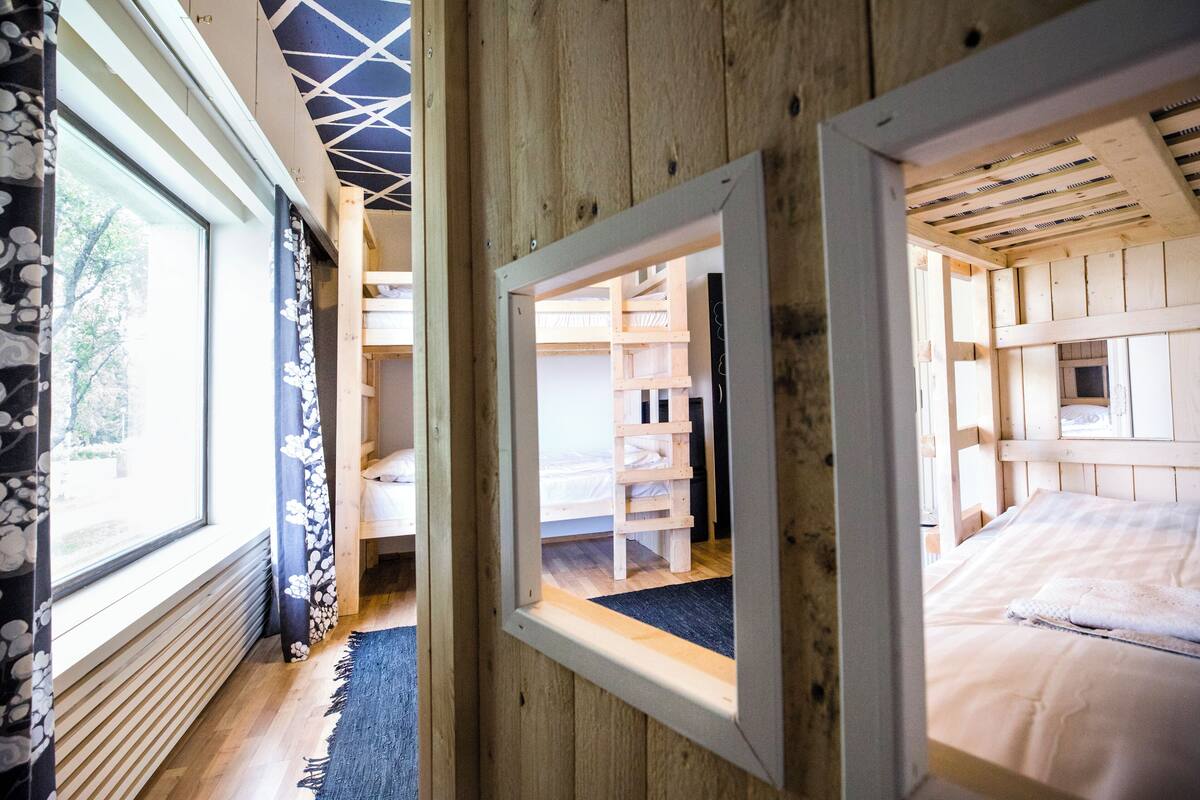 Interior de madera con una litera, una ventana con cortinas y vista a un jardín.