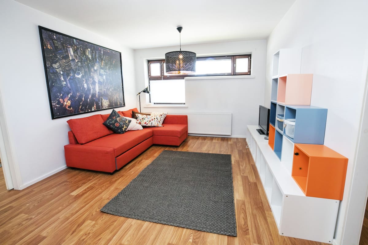 Una sala de estar moderna con un sofá rojo, un cuadro de gran tamaño con una vista urbana, un tapete negro y un estante blanco con bloques de colores.