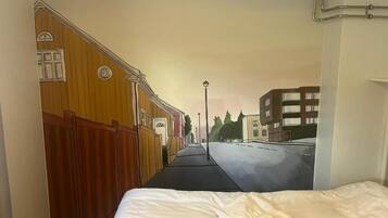 Un dormitorio con una cama, un mural en la pared que muestra una escena callejera y una ventana con persianas.