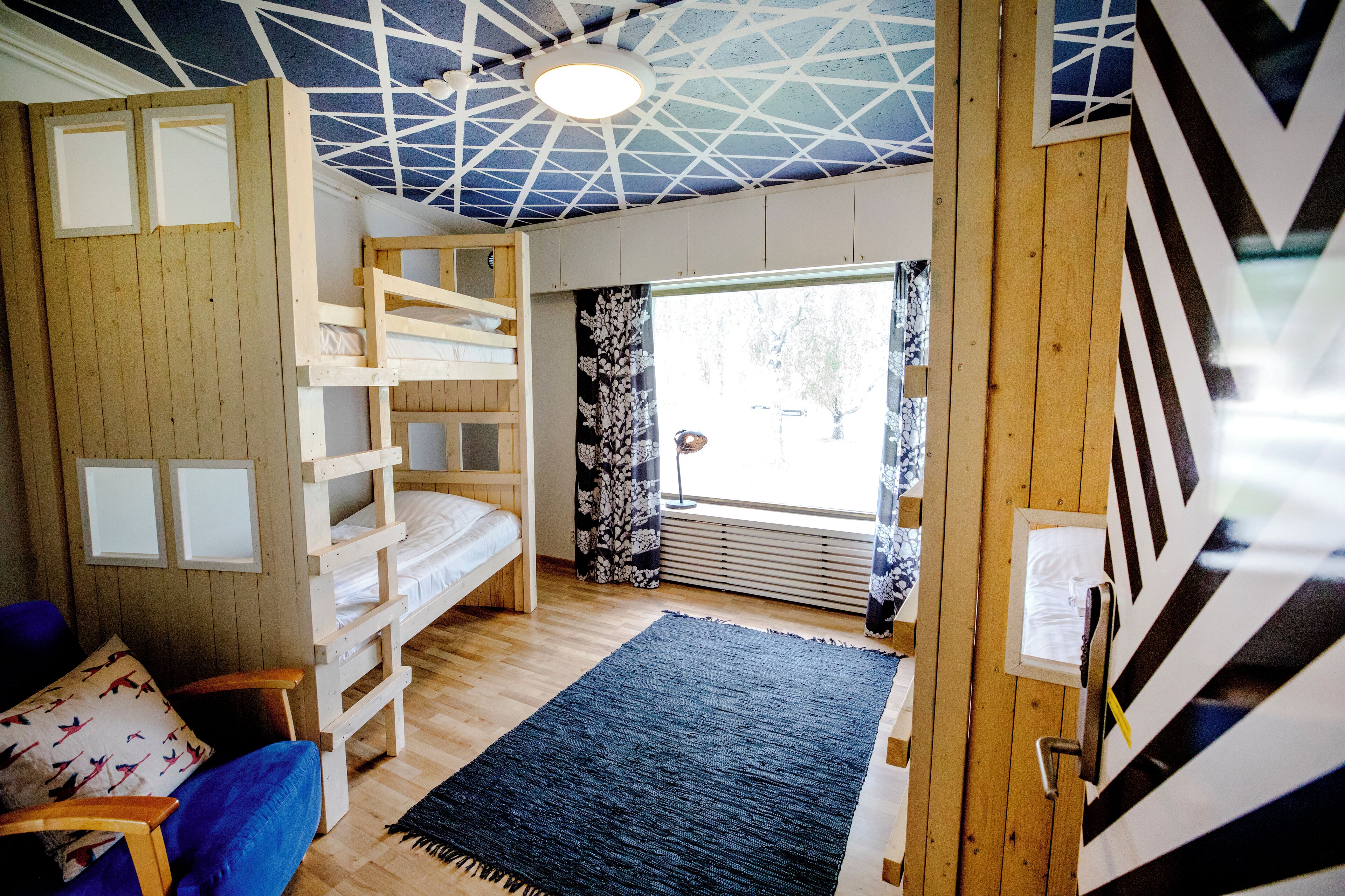 Ett rum med en våningssäng, en blå soffa, en svart matta och ett fönster med utsikt över träd.