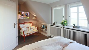 A bedroom with a large bed, a sofa, a desk with a computer, a radiator, and a window with a view. - Ferienhaus Weetstich (Sylt)