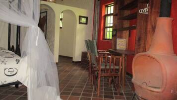 Una habitación con una cama cubierta por una sábana blanca ligera, una mesa de madera con sillas, un jarrón rosa grande y una pared roja con una ventana.