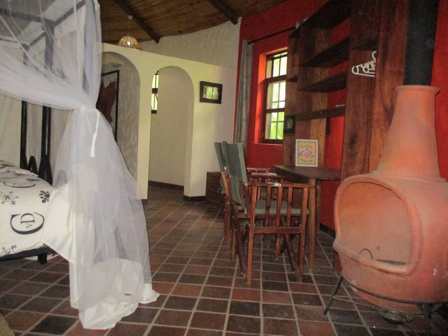 Una habitación con una cama cubierta por una sábana blanca ligera, una mesa de madera con sillas, un jarrón rosa grande y una pared roja con una ventana.