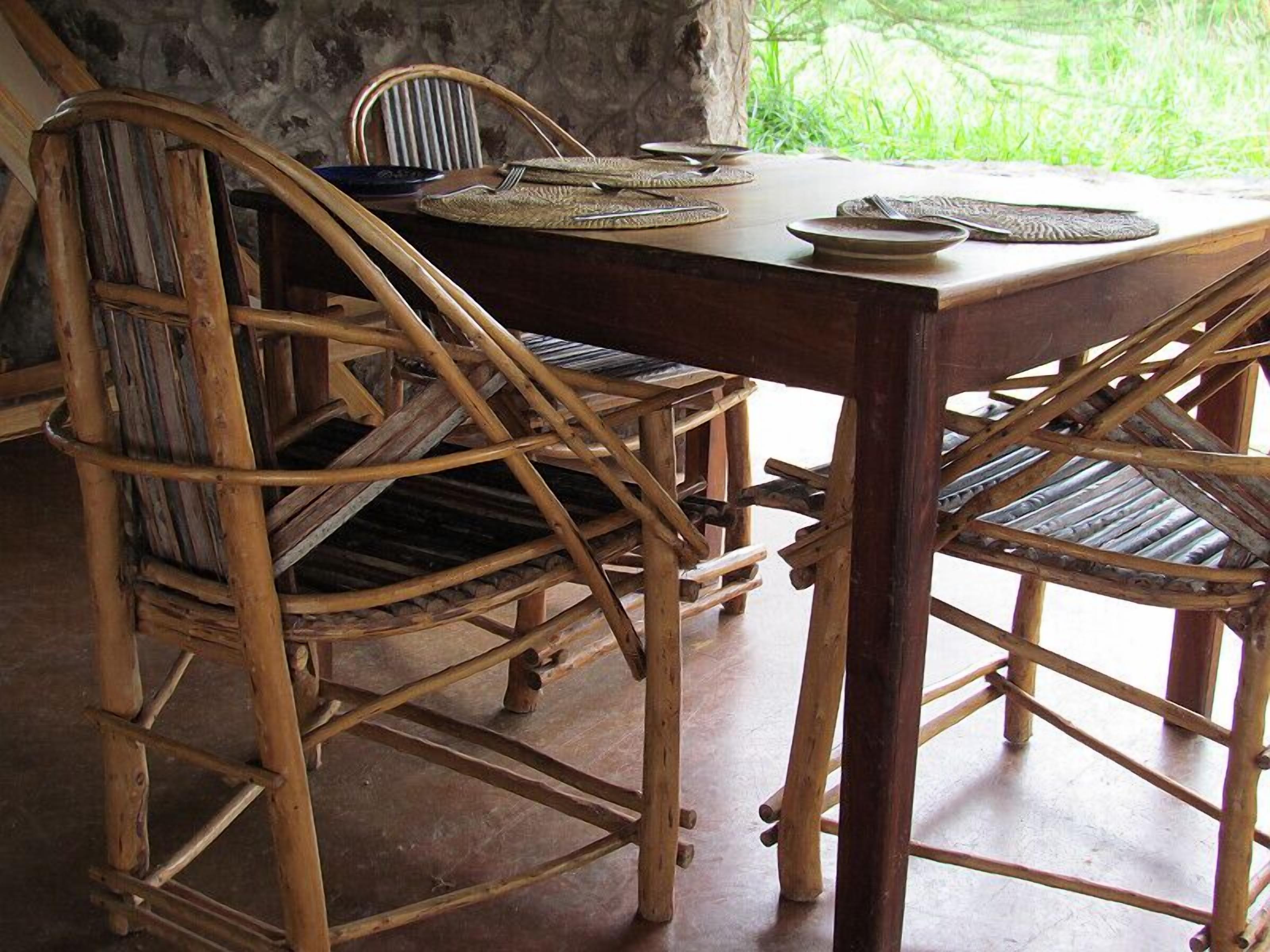 A rustic wooden dining table with woven chairs, a stone wall, and a view of greenery outside.