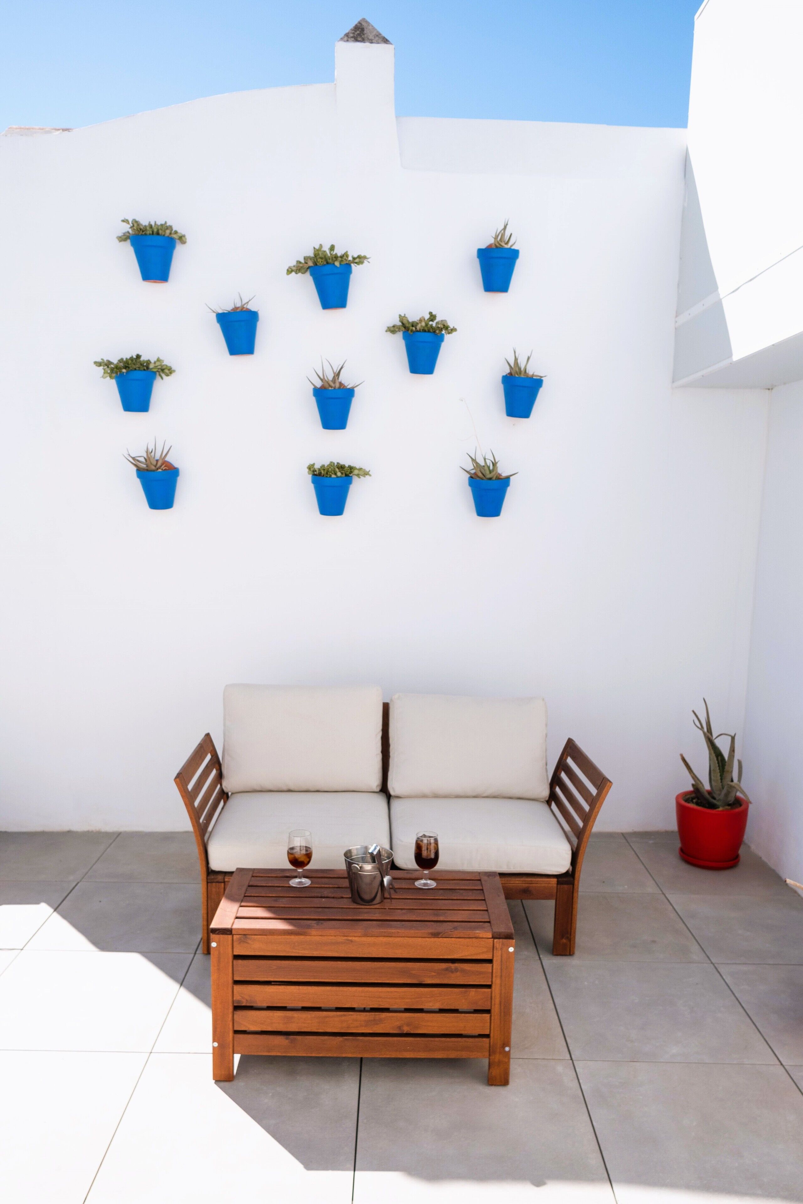 A patio with a wooden table and two chairs, a white wall with blue planters, and a red pot on the floor.
