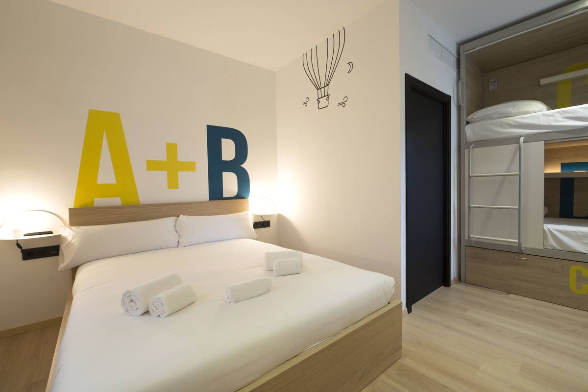 Ein ordentlich gemachtes Bett mit weißen Leinentüchern und zwei aufgerollten Handtüchern, eine Wand mit einem großen 'A+B'-Schild und ein Etagenbett im angrenzenden Raum.
