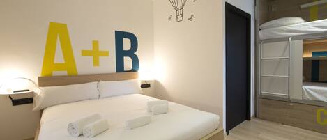 Una cama bien hecha con sábanas blancas y dos toallas enrolladas, una pared con un cartel grande de 'A+B', y una cama nido en la habitación contigua.