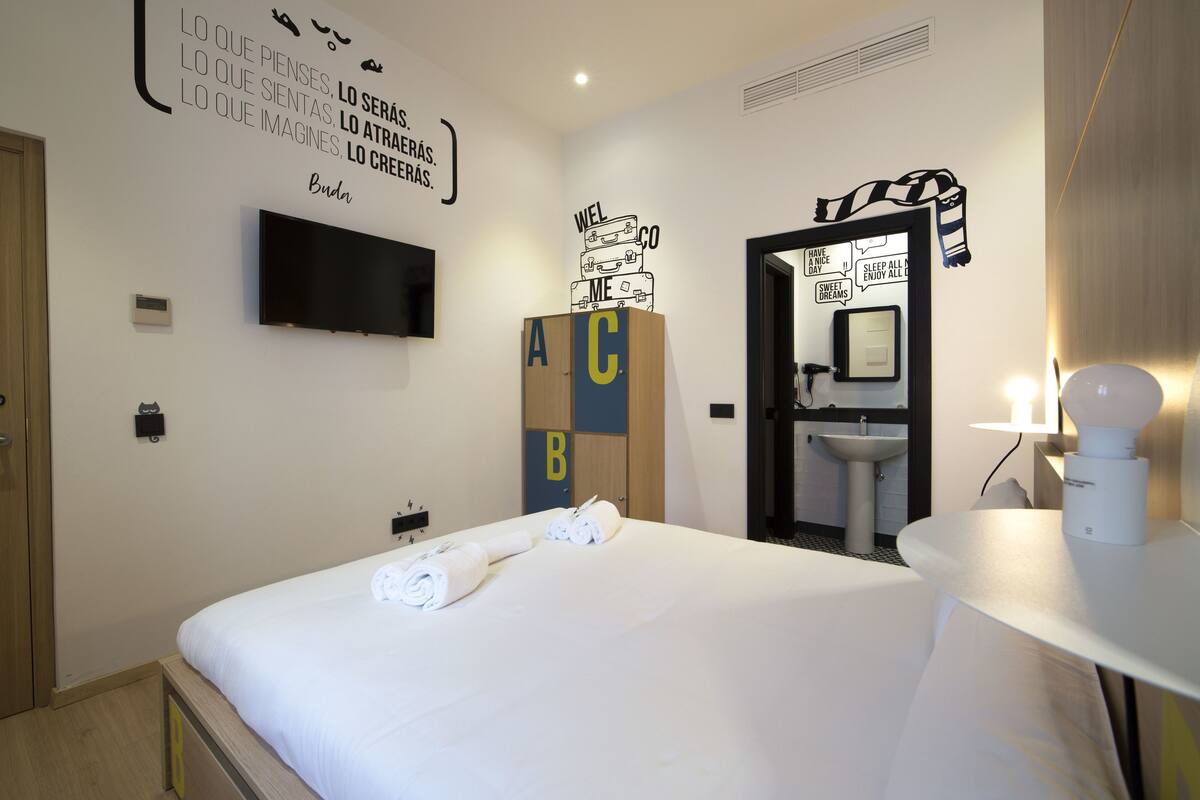 Ein Hotelzimmer mit Bett, einem an der Wand montierten Fernseher, einem Badezimmer mit Waschbecken und motivierendem Wandbild.
