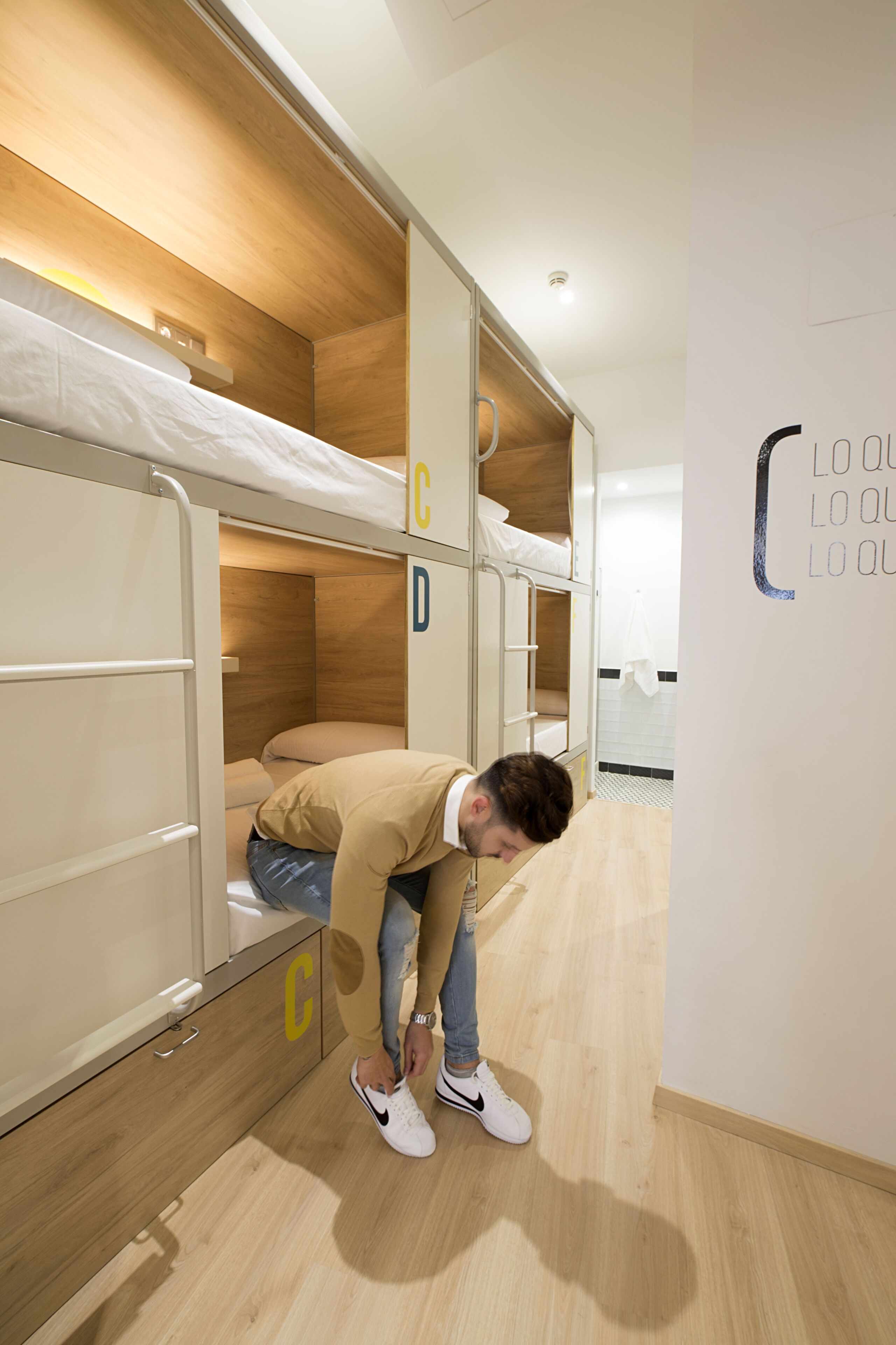 Habitación con literas, decoración de madera, pared blanca con una puerta y una persona ajustándose los zapatos.