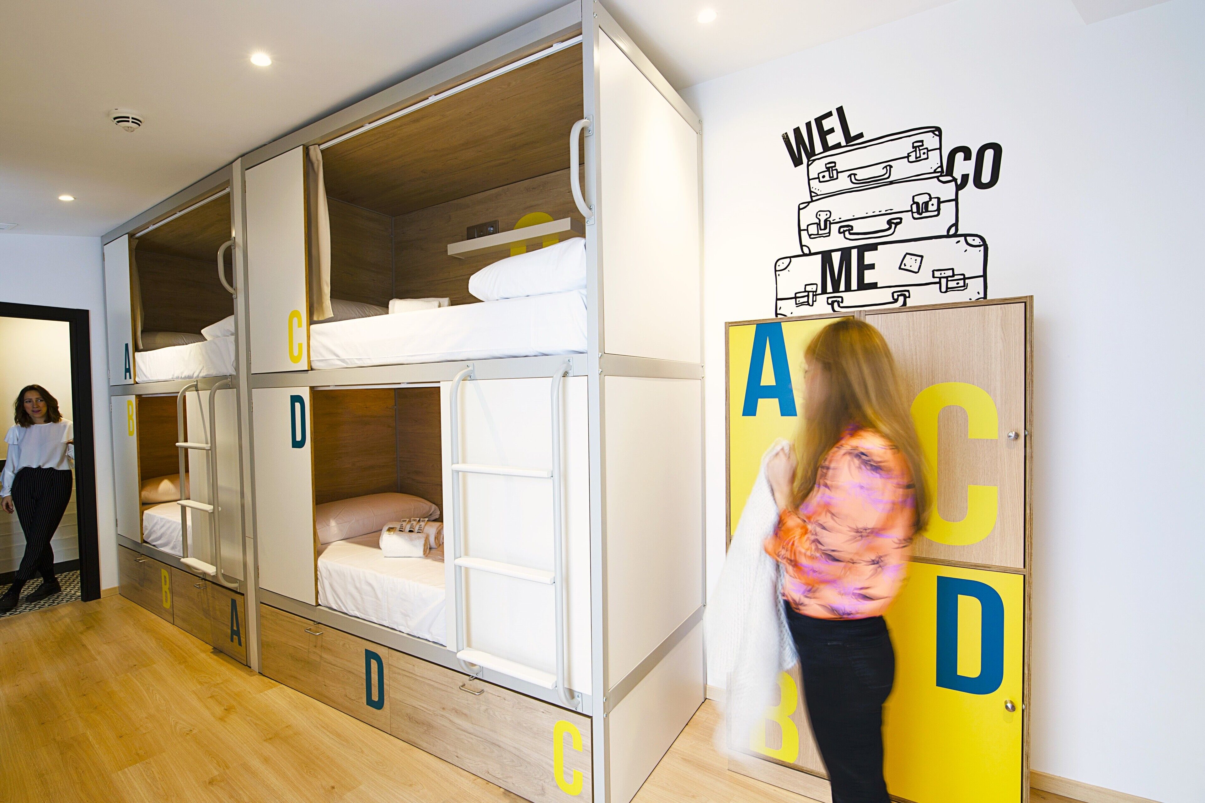 Una habitación con literas identificadas con las letras A, B, C y D, y una pared con un cartel de bienvenida.