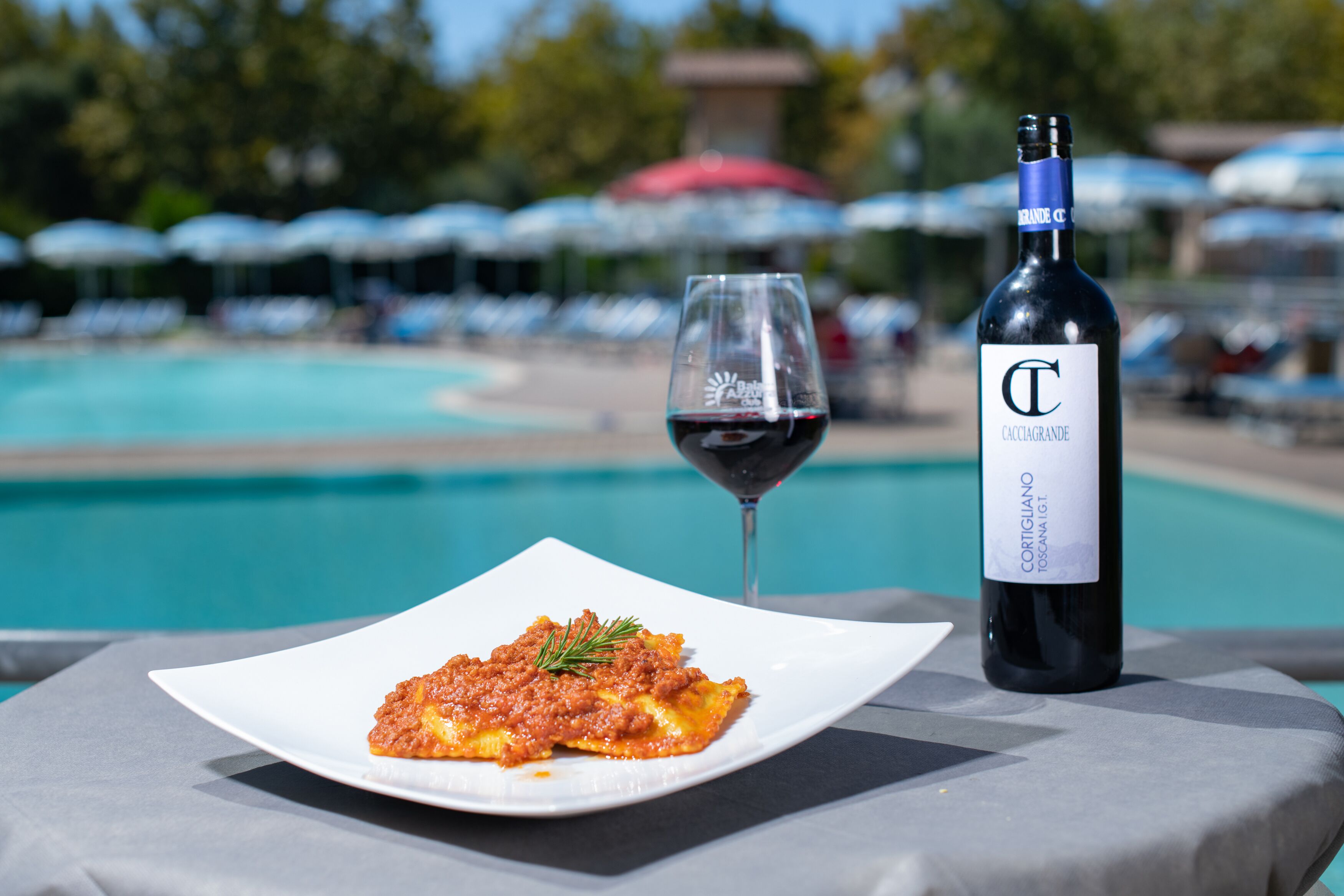 A dining table with a plate of pasta and a bottle of wine, set against a poolside view.