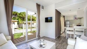 A modern living room with a large sliding glass door to an outdoor deck, a flat-screen TV mounted on the wall, and a dining area with white furniture.