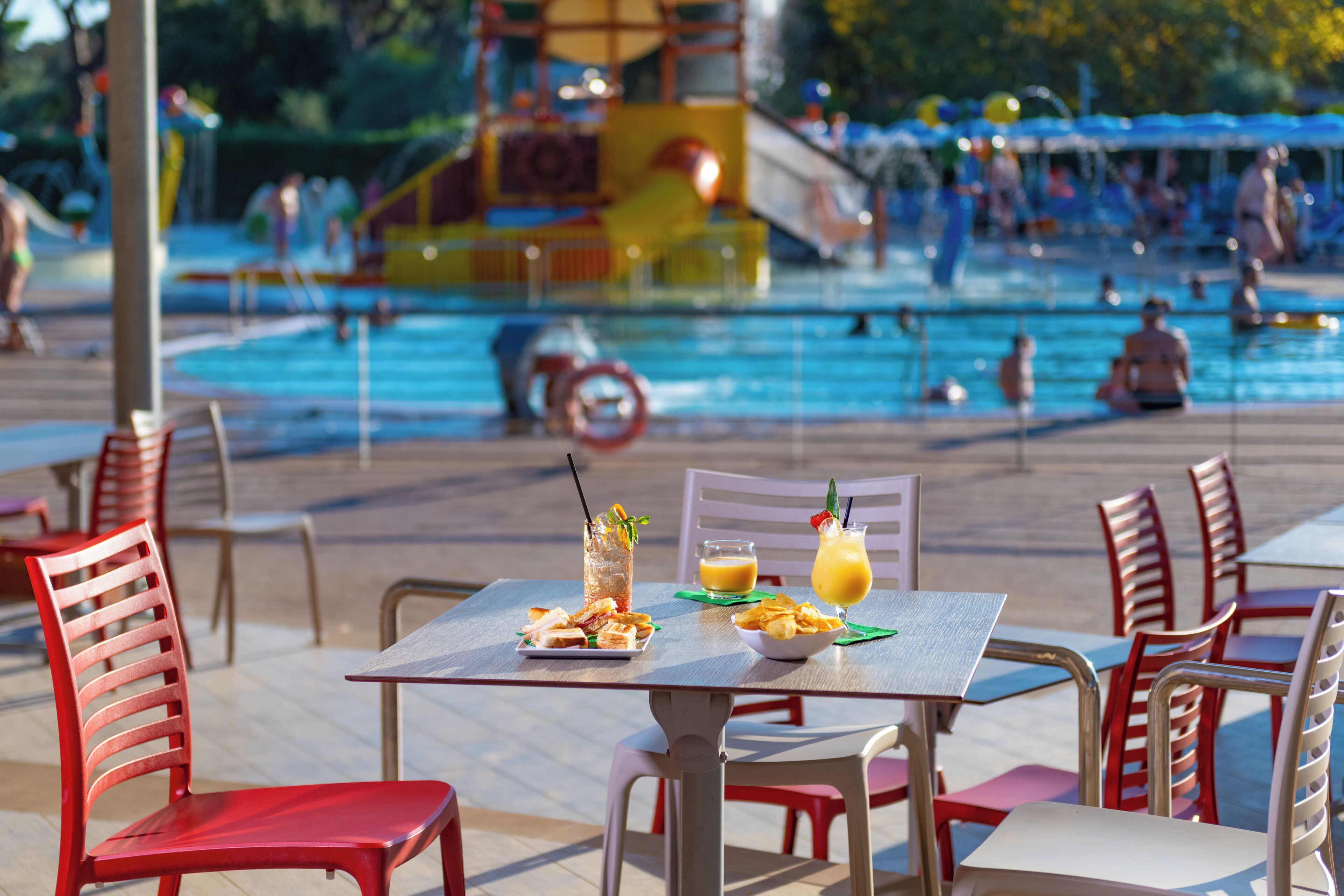 Un espace repas aménagé au bord de la piscine, avec des tables et des chaises, une piscine et un toboggan aquatique en arrière-plan.