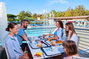 Un grupo de personas cenando en una mesa con vista a la piscina, incluyendo alimentos para el desayuno y un menú.