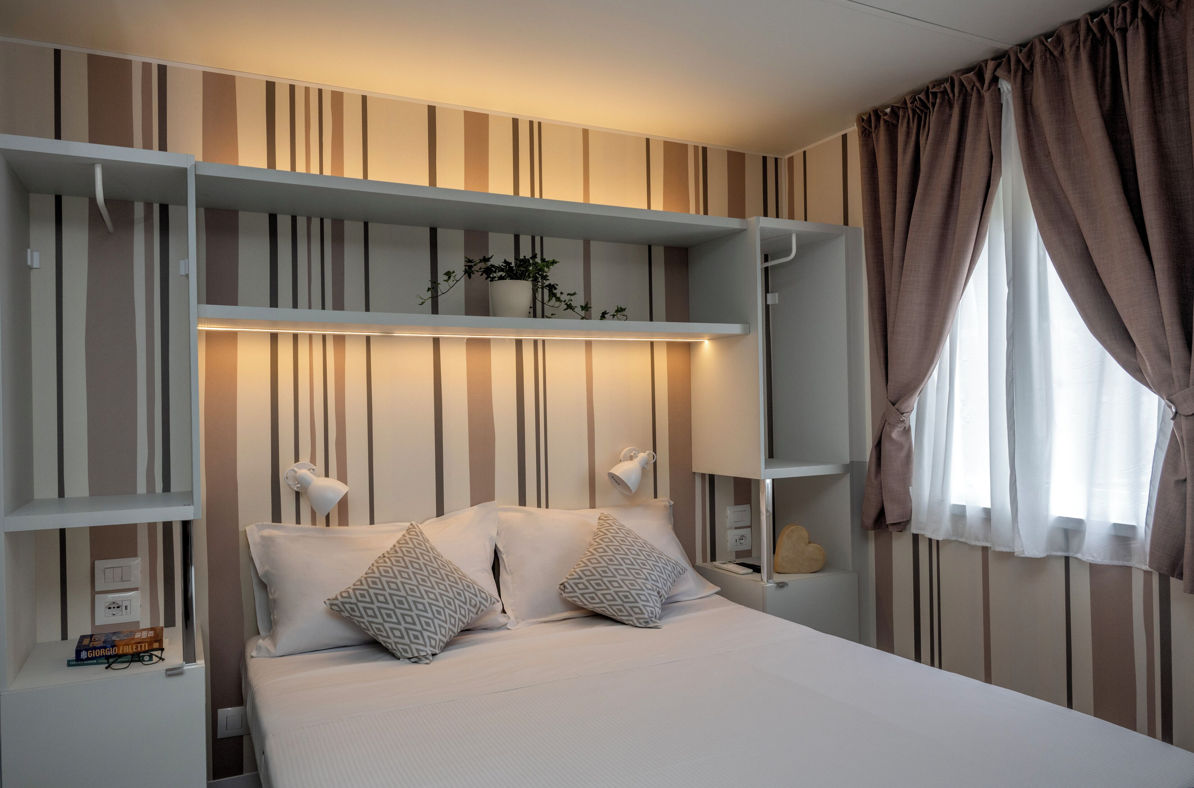 Un dormitorio con cama, dos mesitas de noche, una mesita auxiliar, una estantería con una planta y una ventana con cortinas.