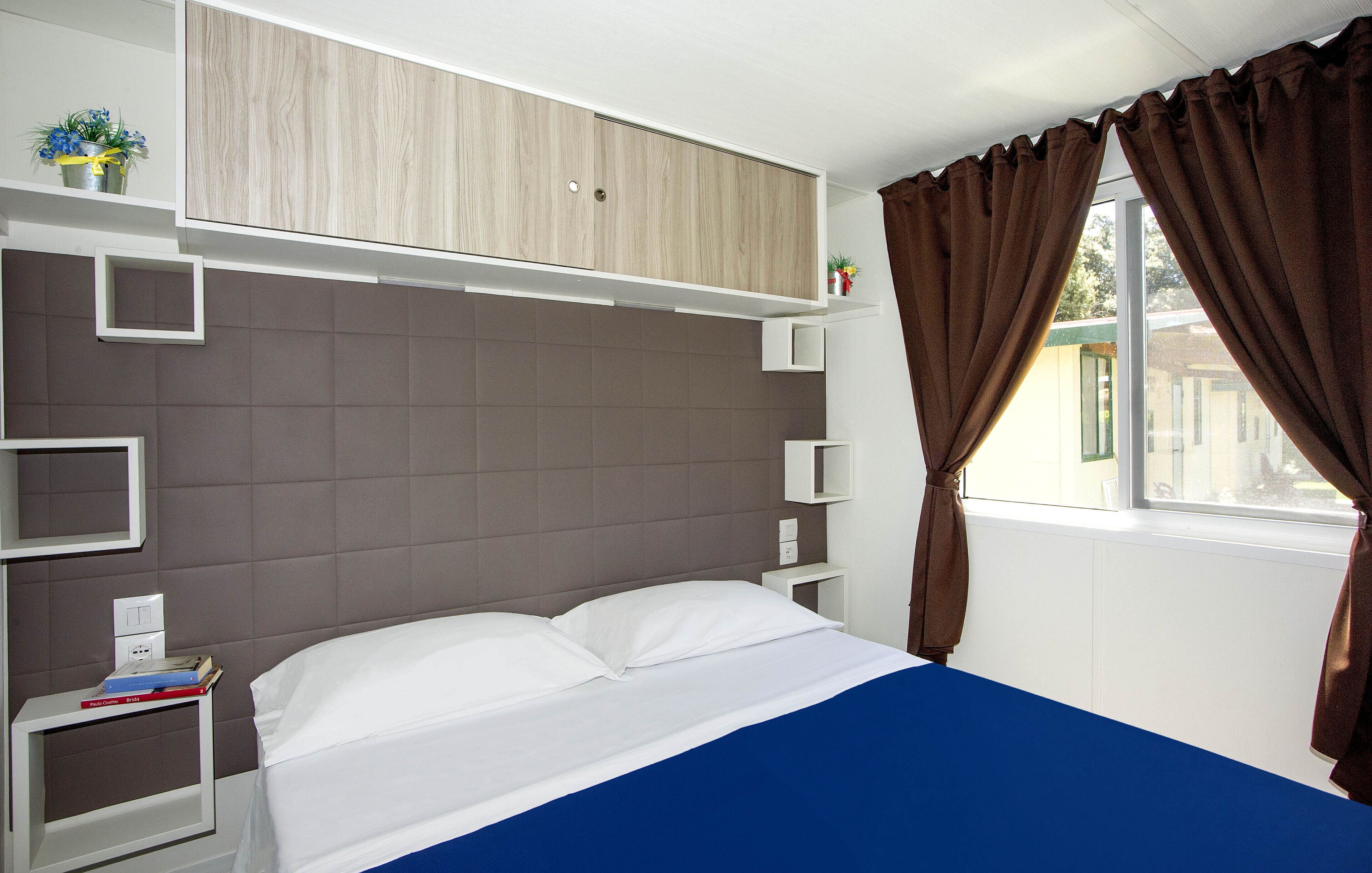 Un dormitorio con cama, mesita de noche, armario y ventana con cortinas.