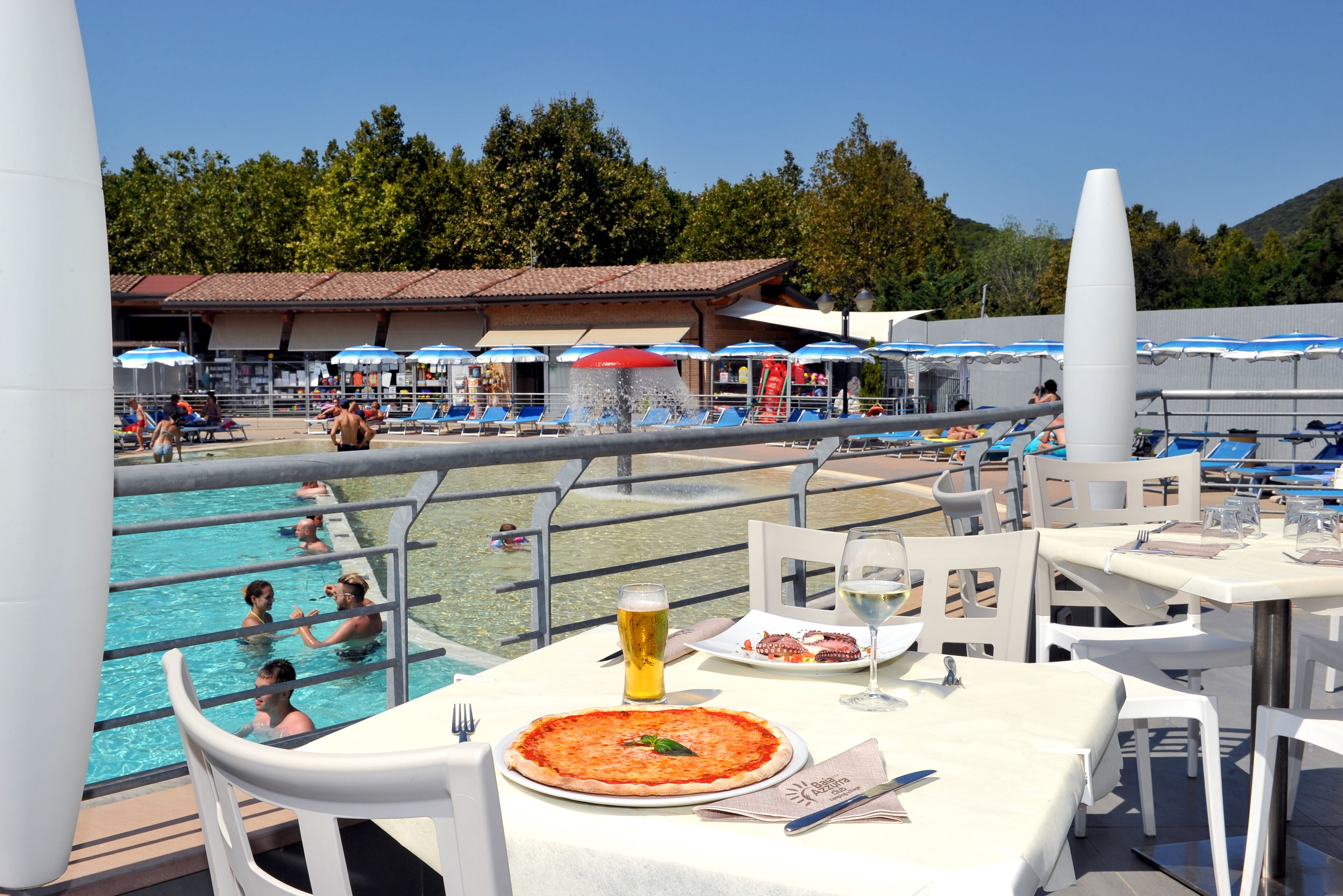 Un área de comedor junto a la piscina con una mesa lista para una comida, una pizza y una cerveza.