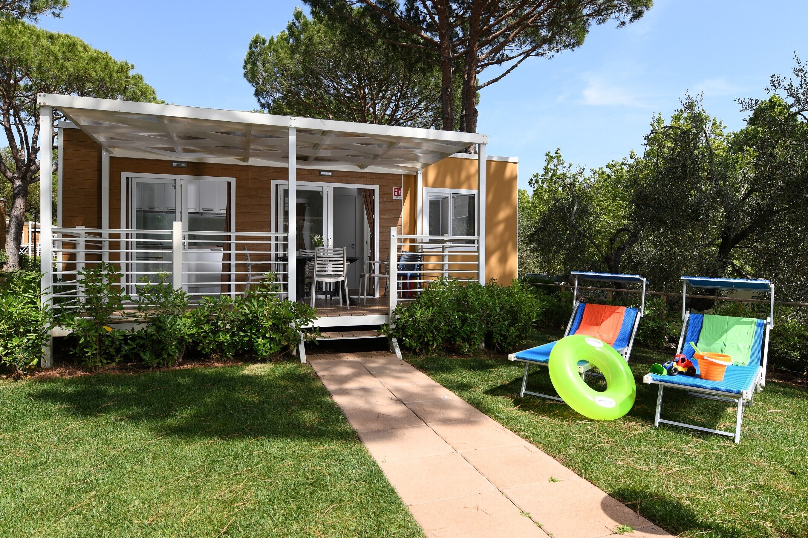 A small, single-story building with a covered porch and a pathway leading to it. There are two lounge chairs with a green inflatable ring and a small table with toys.