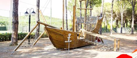 A wooden pirate ship play structure in a park with trees and a fence in the background.