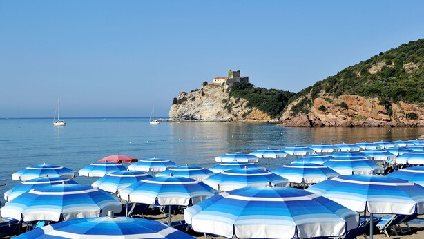 A beach with numerous blue and white striped umbrellas, a castle on a cliff, and calm water with boats.