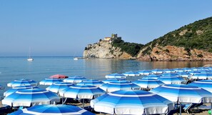 A beach with numerous blue and white striped umbrellas, a castle on a cliff, and calm water with boats. - Camping Village Baia Azzurra (Castiglione della Pescaia)