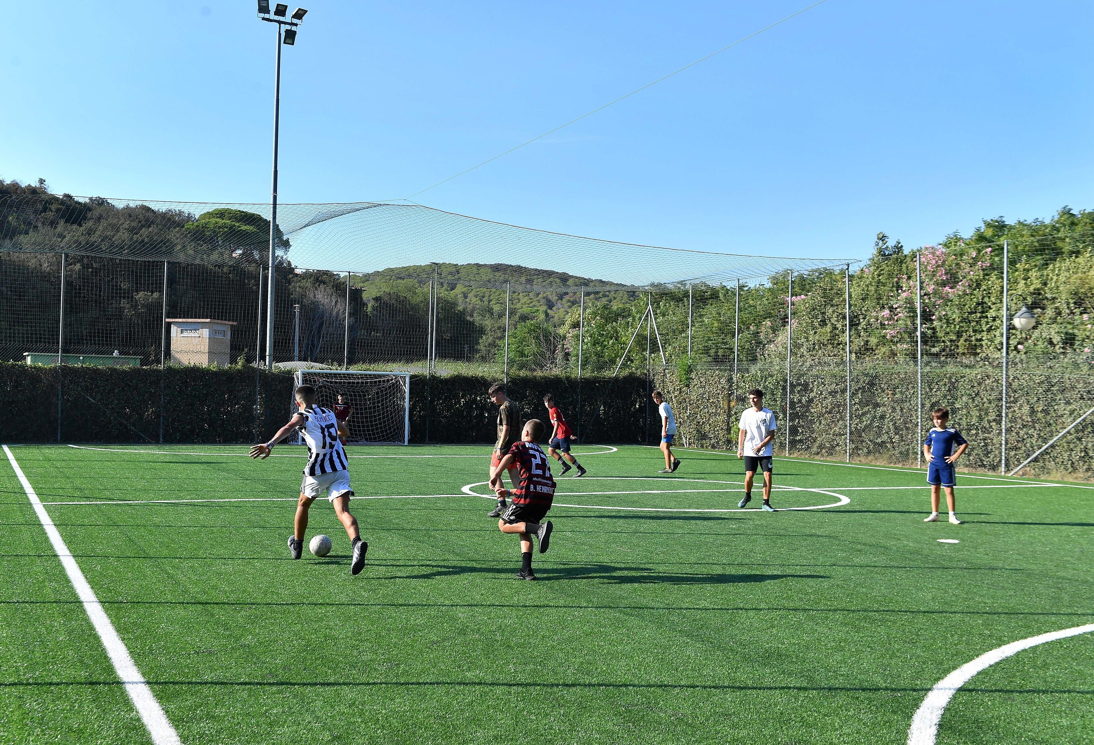 Ein Fußballfeld mit spielenden Spielern, umgeben von einem Zaun und Bäumen im Hintergrund.