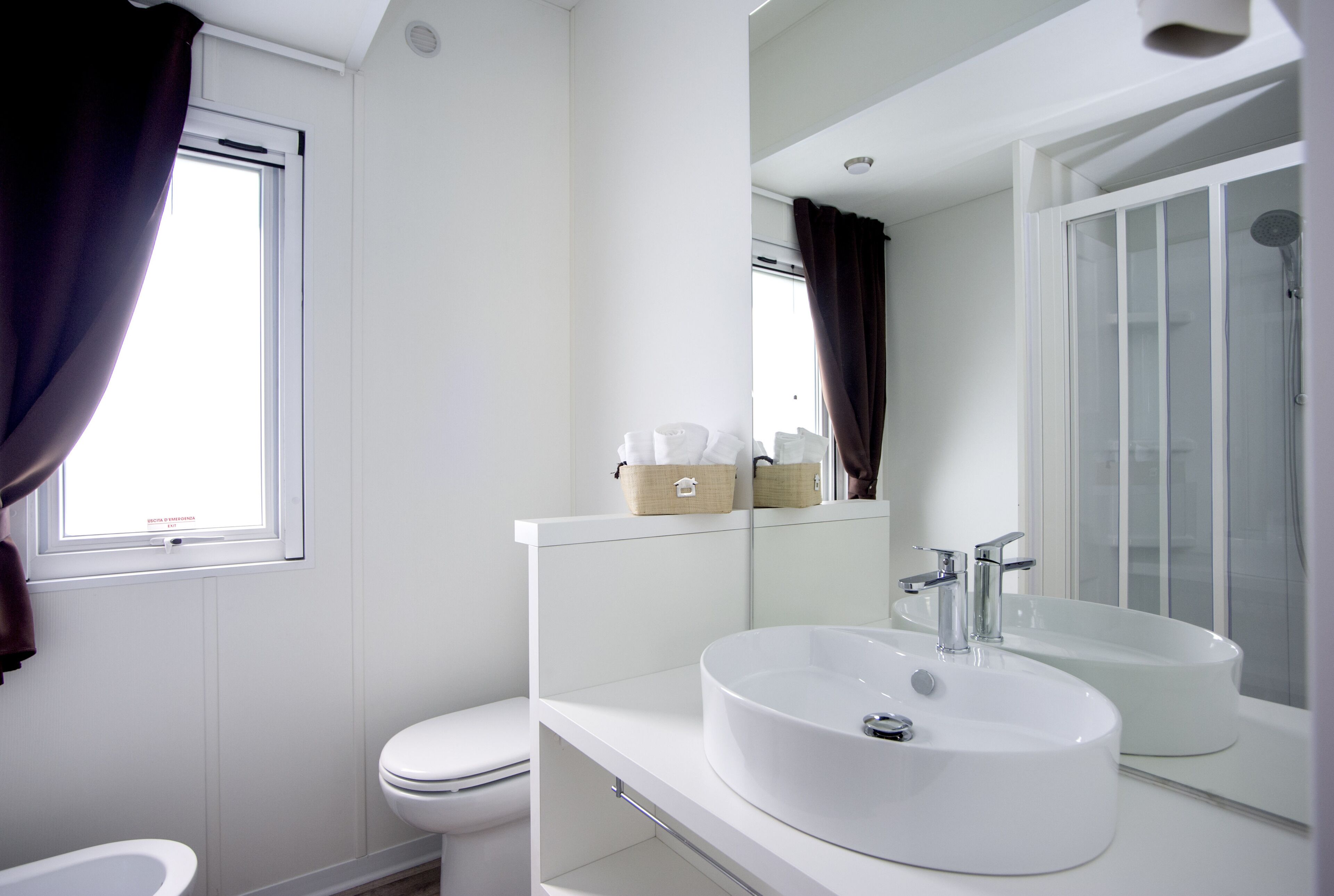 Een moderne badkamer met een wit wastafelmeubel, toilet en douche. Er is een raam met gordijnen, en een spiegel boven het wastafelmeubel.