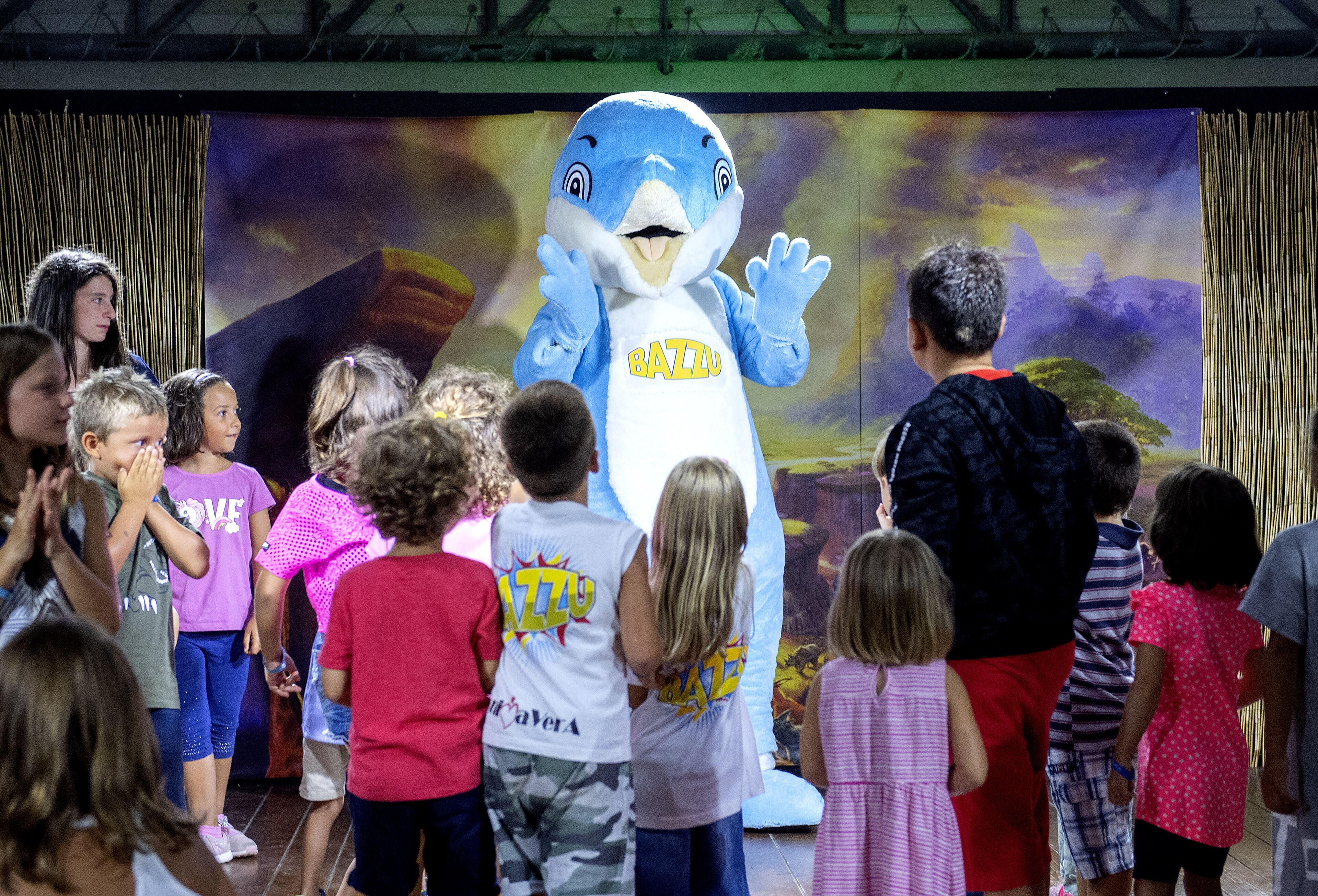 Een mascotte die met kinderen op een podium staat, met een landschap als decor.
